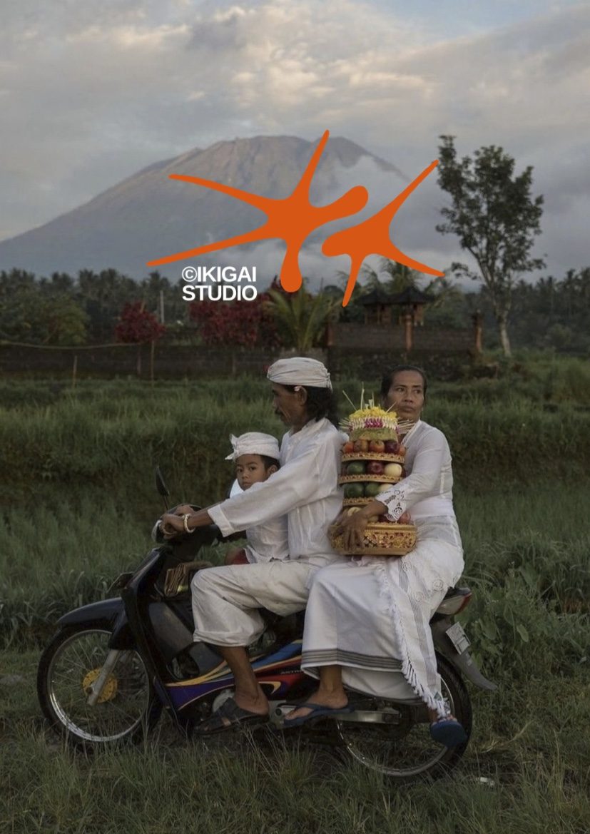 Family in white ceremony dress riding a scooter past rice fields with an offering tower, Mount Agung behind